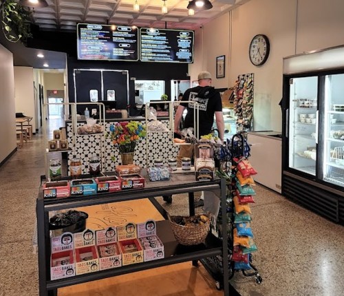 A cozy shop interior with a display of snacks, drinks, and a customer browsing near a menu board and a clock.