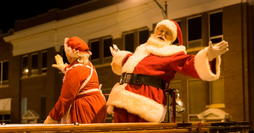 Santa Claus and Mrs. Claus wave joyfully from a festive parade float at night.