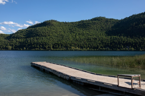 A wooden dock extends into a calm lake, surrounded by lush green hills under a clear blue sky.
