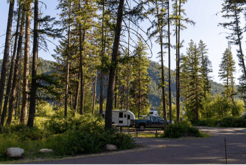 A serene campsite surrounded by tall trees and mountains, featuring an RV parked on a gravel road.
