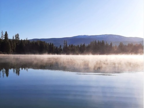 A serene lake at dawn, with mist rising over the water and mountains in the background under a clear blue sky.
