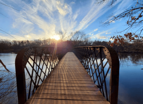 A wooden bridge over a calm river at sunset, with vibrant clouds and trees in the background.