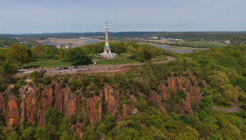 Aerial view of a monument atop a rocky cliff, surrounded by lush greenery and a scenic landscape in the background.
