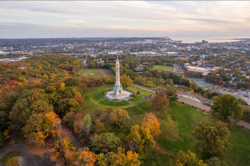 Aerial view of a monument surrounded by colorful autumn trees, overlooking a city and coastline in the distance.