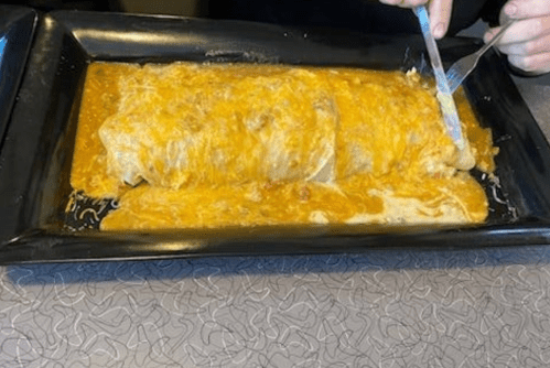 A large burrito covered in a creamy sauce on a black platter, with a person cutting into it.