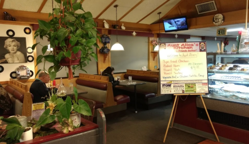 A cozy diner interior with a menu board, plants, and a customer sitting at a booth.