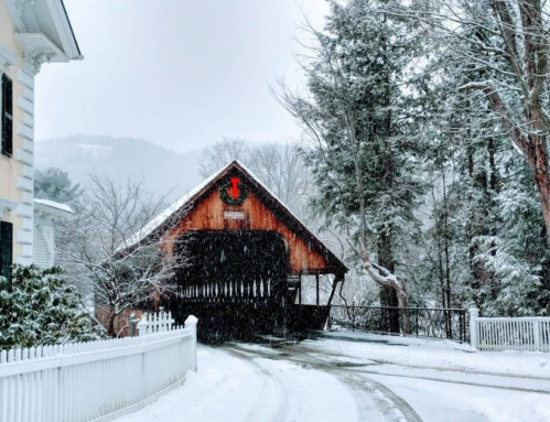 A snow-covered scene featuring a rustic covered bridge surrounded by trees and a white picket fence.