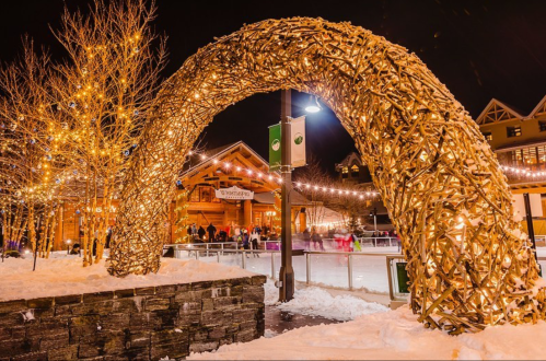 A lit arch made of branches stands near an ice rink, surrounded by snow and festive lights at night.