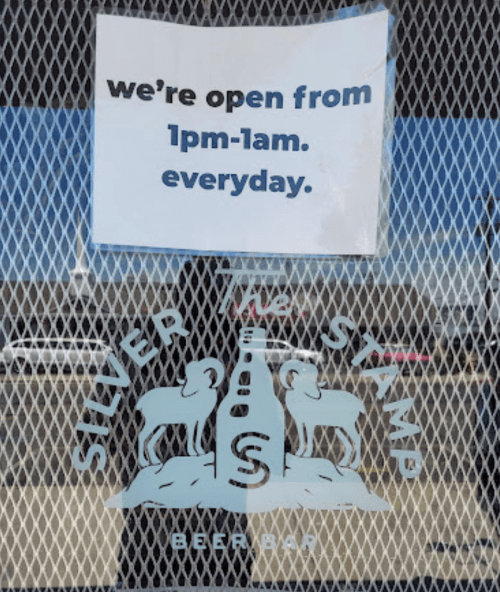 Sign on a window stating "We're open from 1pm-1am every day" at Silver Stamp Beer Bar, featuring a logo with two rams.