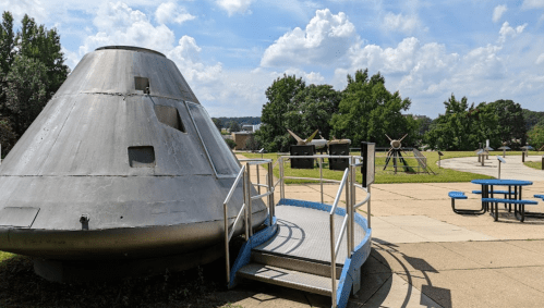 A metallic spacecraft model with a ramp, surrounded by trees and a picnic area under a blue sky with clouds.