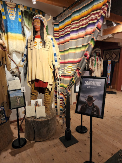 A Native American figure stands beside colorful textiles and a poster for "Longmire" in a museum display.