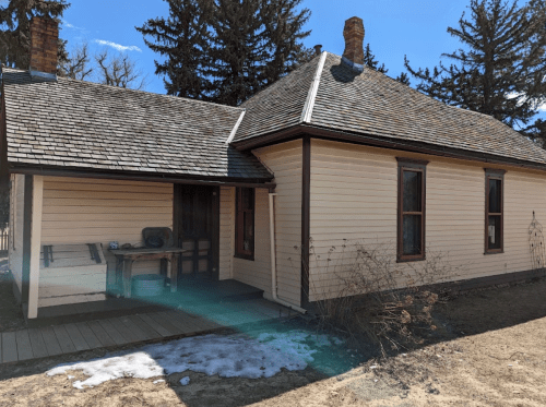 A historic wooden house with a sloped roof, surrounded by trees and patches of snow on the ground.