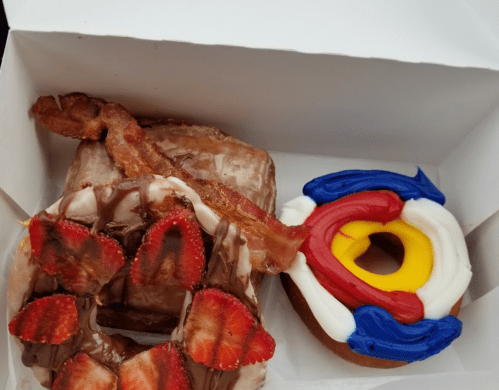 Two donuts in a box: one topped with chocolate, strawberries, and bacon; the other decorated with colorful icing.