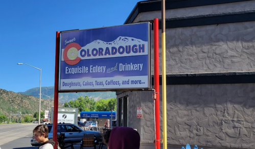 Sign for "Sweet Coloradough," an eatery featuring doughnuts, cakes, teas, and coffees, with mountains in the background.