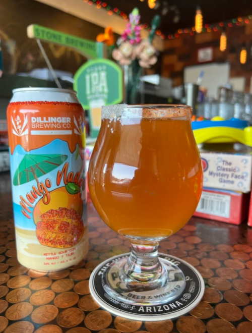 A glass of amber beer sits on a coaster next to a can of Mango Nachos beer, with a colorful bar background.