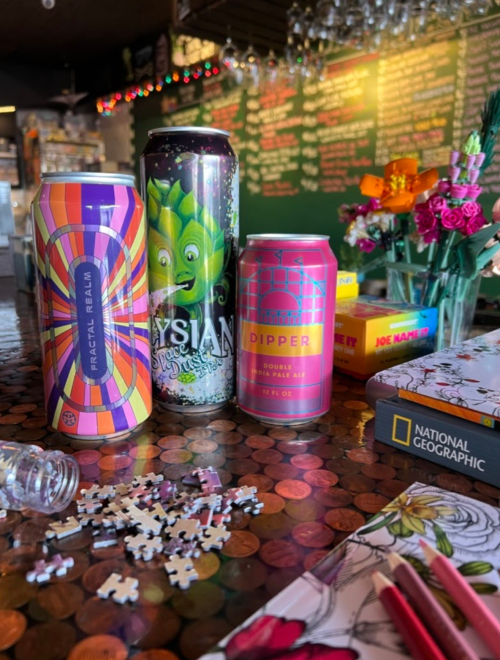 Three colorful beer cans on a table with a puzzle, flowers, and a National Geographic magazine in a cozy setting.
