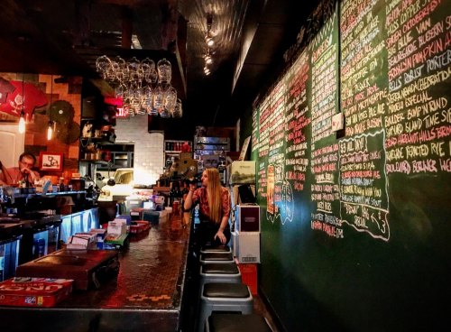 A cozy bar with a chalkboard menu, shelves of glasses, and a woman sitting at the counter, enjoying the atmosphere.