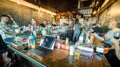 A lively bar scene with patrons seated at the counter, games on the table, and a chalkboard menu in the background.