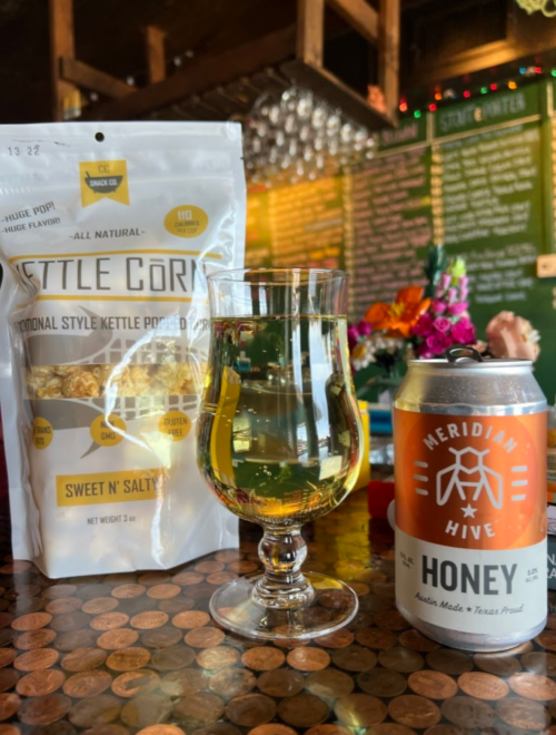 A glass of honey-colored drink next to a bag of kettle corn and a can of honey beverage on a table with coins.