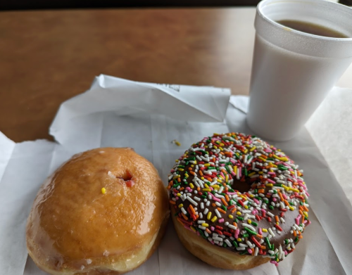 A glazed donut and a chocolate frosted donut with sprinkles, next to a cup of coffee on a paper wrapper.