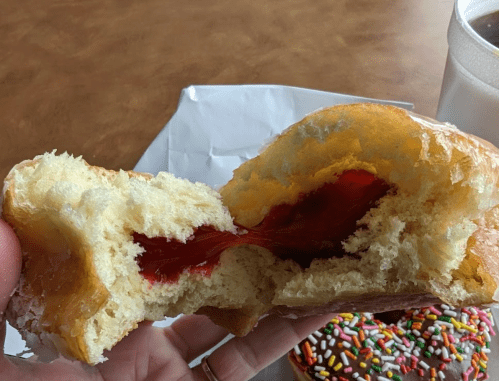 A hand holding a split donut revealing a red jelly filling, with a chocolate donut and drink in the background.