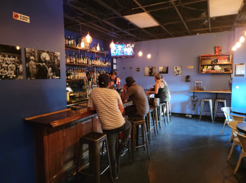 A cozy bar with patrons seated at the counter, surrounded by shelves of liquor and blue walls, with a TV in the background.