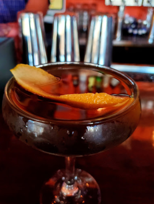 A cocktail in a coupe glass garnished with a lemon twist, set on a wooden bar with metal shakers in the background.