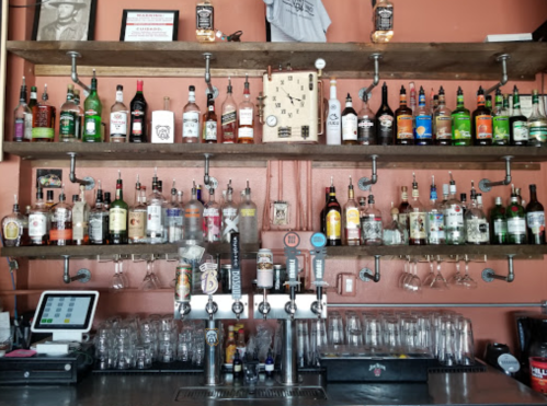 A bar shelf displaying various bottles of liquor and a beer tap setup, with glasses and a register in the foreground.