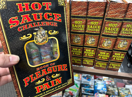 A person holds a box labeled "Hot Sauce Challenge: Book of Pleasure & Pain," with bottles of hot sauce visible inside.