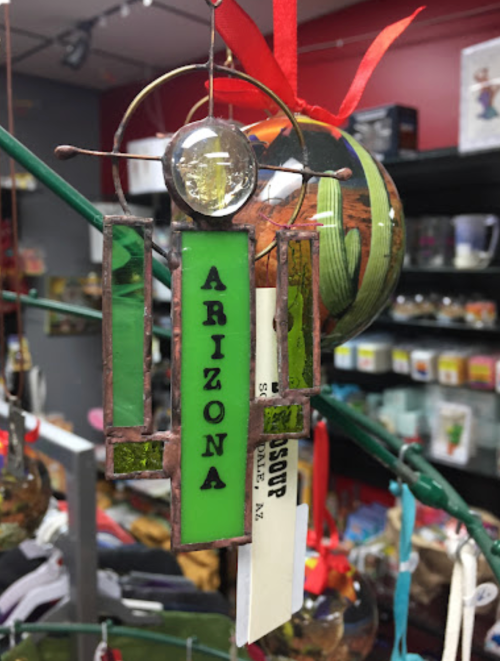 A colorful ornament featuring the word "Arizona" in green glass, hanging with a red ribbon in a shop setting.