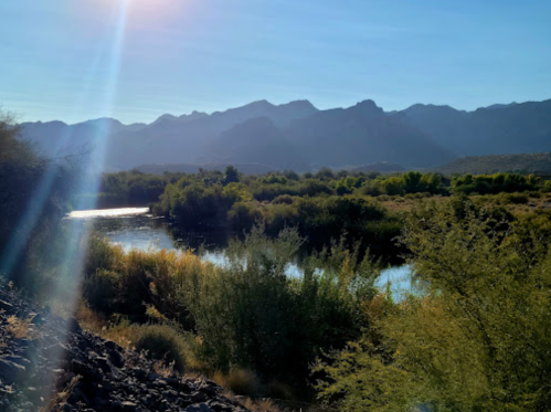 A serene river flows through a lush landscape, framed by mountains under a clear blue sky. Sunlight glimmers on the water.