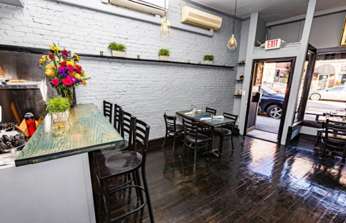 Cozy restaurant interior with wooden tables, black chairs, and colorful flower arrangements against a brick wall.