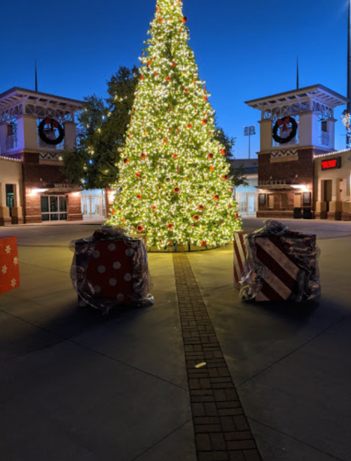 A brightly lit Christmas tree stands in a plaza, flanked by wrapped gift boxes, under a twilight sky.