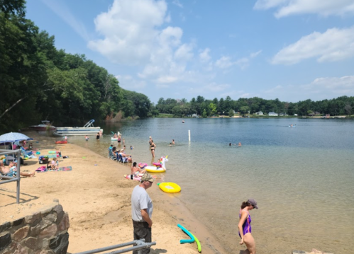 A sunny lakeside scene with people swimming, sunbathing, and enjoying the beach on a warm day.