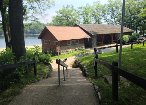 A pathway leads down to a lakeside building surrounded by trees and greenery, with a beach in the background.
