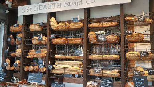 A display of various artisanal breads in baskets at Olde Hearth Bread Co., with price tags visible.