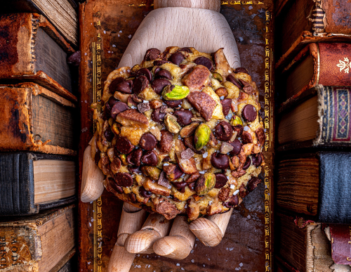 A wooden hand holds a large cookie topped with chocolate chips and nuts, surrounded by old books.
