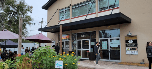 A modern building with large windows, outdoor seating, and people walking by on a cloudy day.