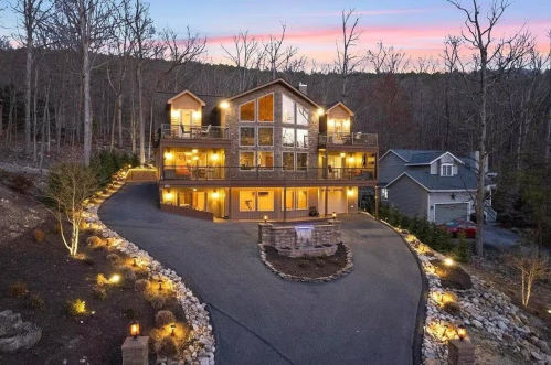 A large, modern house with multiple balconies, surrounded by trees and illuminated by outdoor lights at dusk.