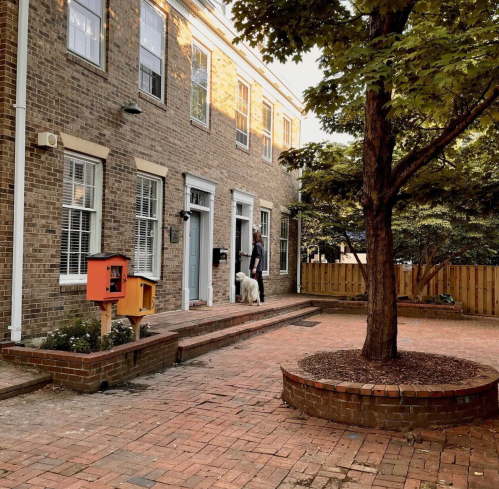 A person stands at the door of a brick house with a dog, surrounded by a landscaped yard and a tree.