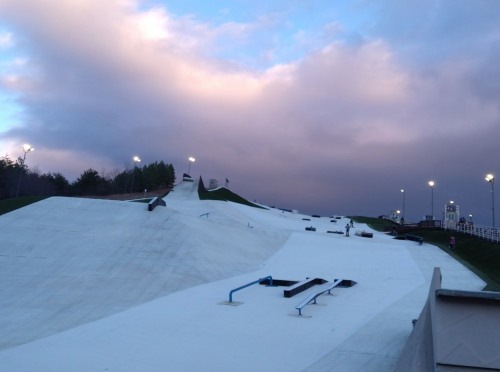 A snowboarding slope with ramps and rails under a cloudy sky, illuminated by lights in the background.
