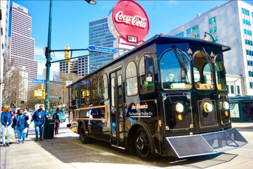 A vintage-style trolley bus parked on a city street near a Coca-Cola sign, with pedestrians walking nearby.