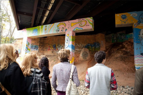A group of people observes colorful murals on pillars under a bridge, surrounded by graffiti and natural scenery.