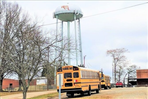 A yellow school bus parked near a water tower with a tiger logo, surrounded by trees and a cloudy sky.