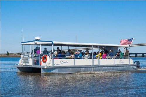 A tour boat named "Liberty" with passengers, cruising on a calm river under a clear blue sky. An American flag is visible.