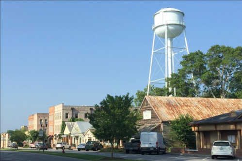 A small town scene featuring a water tower, historic buildings, and trees under a clear blue sky.