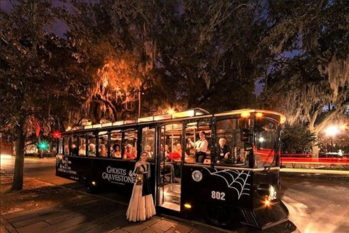 A spooky trolley bus with passengers, parked under trees draped in Spanish moss at night, promoting a ghost tour.