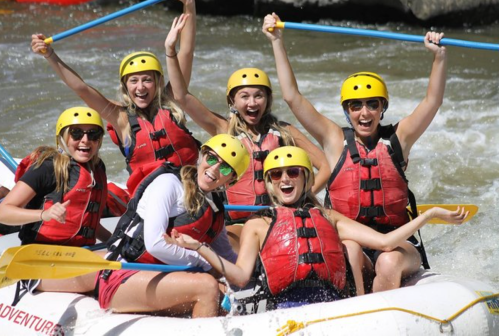 A group of seven women in yellow helmets and life jackets joyfully pose in a raft on a river, surrounded by splashing water.