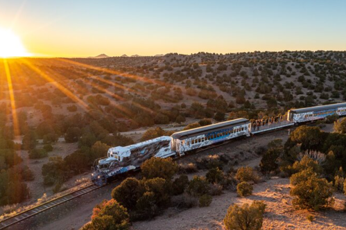 A colorful train travels along a scenic landscape at sunset, with sun rays illuminating the surrounding hills.