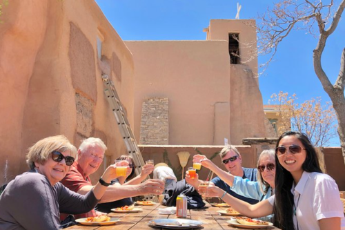 A group of six people at an outdoor table, raising glasses in a toast, with a rustic building in the background.